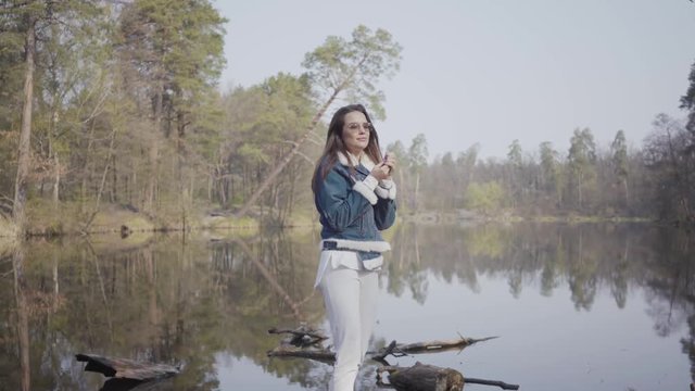 Pretty Young Woman In White Pants, Jeans Jacket And Sunglasses Standing On The Riverbank. The Girl Is Cold, She Rubbing Her Hands Trying To Warm Herself. Beautiful Landscape On Background