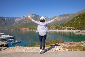 Fototapeta na wymiar Stylishly dressed girl in blue jeans, white hat, shirt and sneakers enjoys traveling during her spring vacation in the Kiparissi Lakonia village, Peloponnese, Zorakas Bay, Greece, May 2019.
