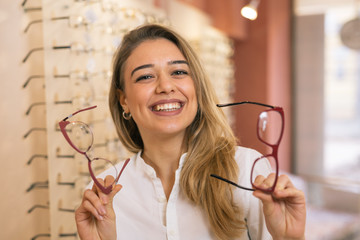 pretty smiling woman choosing eye glasses in optic store - obrazy, fototapety, plakaty