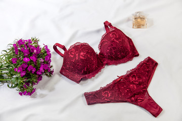 Red women's bra and panties on white bed background. Concept photo decorated with purple colored flowers and perfume. Underwear fashion. Basic lingerie. Classic bra - Image