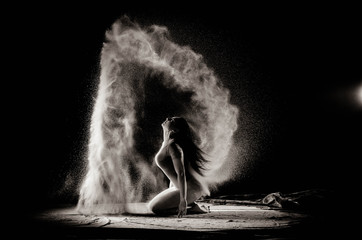 The girl with the flour on the body stretches the arms up with thrown flour on black background...
