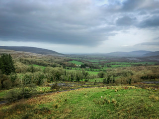 Fototapeta na wymiar Landscape Shot Overlooking the Countryside in Southern Ireland - Taken Above a Long, Winding Road, with Pastures, Mountains and a Cloudy Sky in the Background