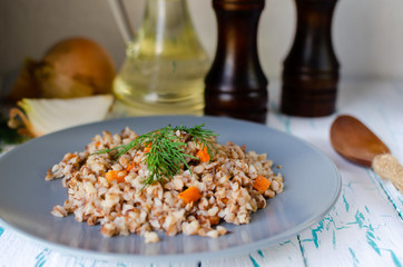 Buckwheat porridge with carrots and dill in a blue plate. On background of Golden onions, green dill and salt and pepper mills.