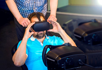 Fototapeta na wymiar Mature bearded man wears VR glasses. Gamer plays VR race on a dark room gaming club. Virtual reality on car simulator. A man is sitting behind a car simulator and plays games.