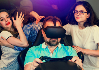 Mature father is playing racing videogame in 3D virtual reality simulator using headset. Big family having fun together. Frightened children are looking at the screen. Selective focus