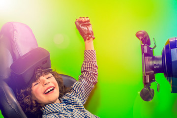 Toddler boy with a virtual reality headset sitting in a autosimulator triumphantly raising his hand up on a green background.technology,entertainment concept - young man playing car racing video game.