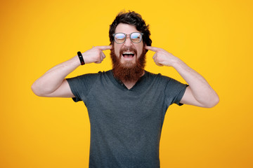 Photo of handsome man with closed eyes, scream and putting finger in his ears, standing over yellow background