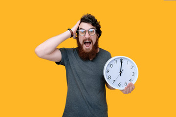 Photo of frustrated bearded guy, holding a clock and looking at camera over yellow background