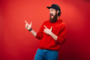 Excited bearded man in red hoody, is pointing away with finger, over isolated background