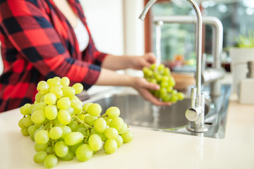 Green grapes on the first plan and a woman rinses  the green grapes with water in the kitchen.
