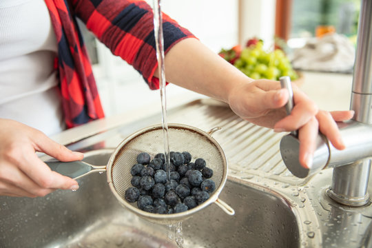 Woman Rinses Blueberries On The Strainer With Water In The Kitchen With Fruit Bowl On The Background.