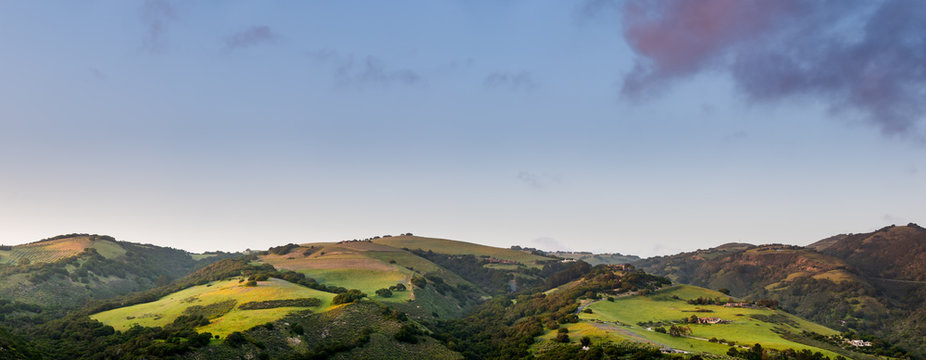 Panorama of beautiful green hills, mountains, and fields in sunset light under a blue sky with purple clouds in Carmel Valley, CA, USA