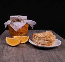 A jar of orange syrup with orange slices next to a plate of fried pancakes on a wooden surface with a black background