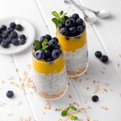 Two Glasses of chia pudding with blueberries, mango mousse and oat granola on the kitchen table.