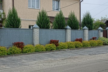 a long gray fence of metal and bricks outside in green grass and ornamental plants outside the road