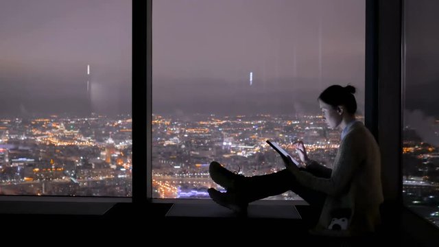 Woman using black digital tablet near window of skyscraper with view night cityscape. Light city street bokeh illumination at evening. Entertainment, opportunity and technology concept