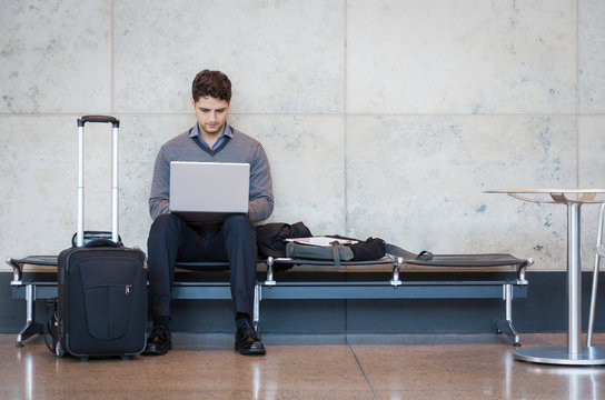 Professional Man Business Traveler With Roller Bag Suitcase Using Laptop Computer In Airport Terminal Waiting Area