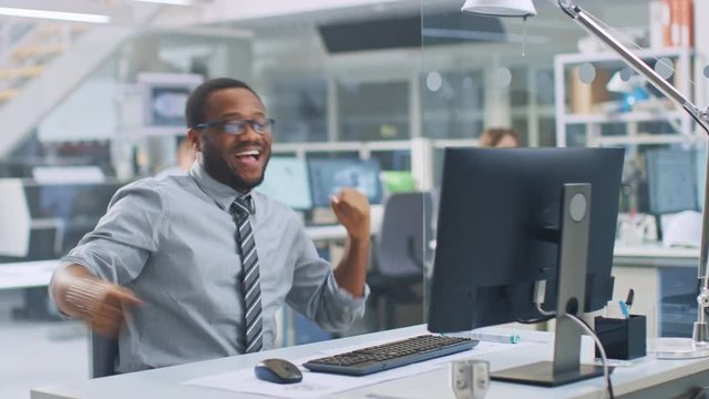 In Industrial Engineering Facility: Portrait of the Smart and Handsome Male Engineer Working on Desktop Computer, Dancing Cheerfully and Celebrating His Success. In the Background Technicians Working