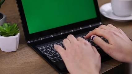 Woman types on laptop pad with green screen. Close up of female hands typing and green tablet on the table.