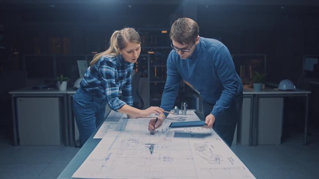 In the Dark Industrial Design Engineering Facility Male and Female Engineers Talk and Work on a Blueprints Using Conference Table. On the Desktop Drawings, Drafts and Electric Engine Components, Parts
