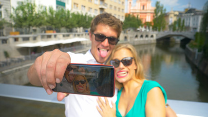 CLOSE UP: Tourist couple making goofy faces while taking selfies from a bridge.