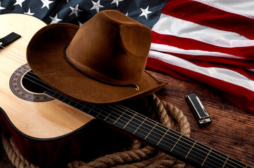American culture, living on a ranch and country muisc concept theme with a cowboy hat, USA flag, acoustic guitar, harmonica and a rope lasso on a wooden background in a old saloon - obrazy, fototapety, plakaty