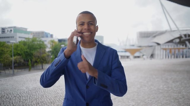Smiling handsome young man talking on phone while walking. Cheerful African American businessman having conversation through smartphone during stroll on square. Technology concept