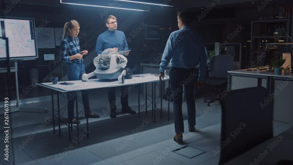 Poster Late at Night in Robotics Engineering Facility Two Technical Engineers Talk and Work on a Wheeled Robot Prototype, Joined by the Chief Engineer. Research Center with Screens Showing Industrial Design