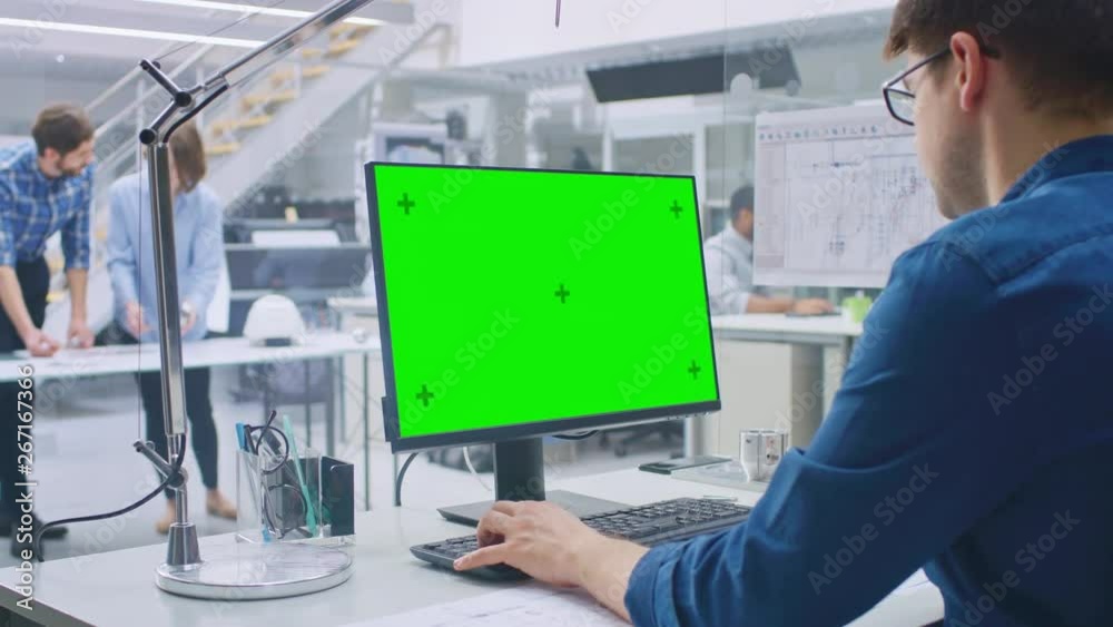 Poster Over the Shoulder Shot of Engineer Working with Green Mock-up Screen Desktop Computer. In the Background Engineering Facility with Specialists Working on Blueprints and Drawings with Industrial Design