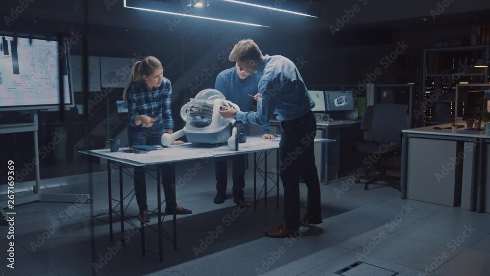 Leinwandbilder Late at Night in Robotics Engineering Facility Three Technical Engineers Talk and Work on a Wheeled Robot Prototype. In the Background High Tech Research Center with Screens Showing Industrial Design