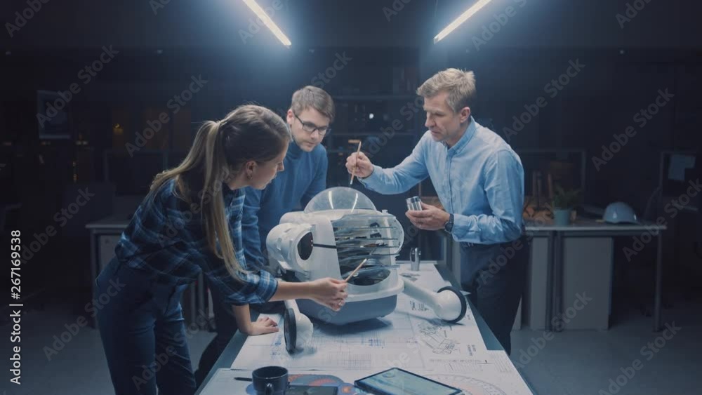 Poster Late at Night in Robotics Engineering Facility Three Technical Engineers Talk and Work on a Wheeled Robot Prototype. In the Background High Tech Research Center with Screens Showing Industrial Design