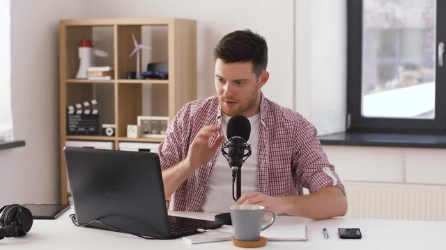 technology, mass media and audioblog concept - happy young man with laptop computer and notebook speaking to microphone at home