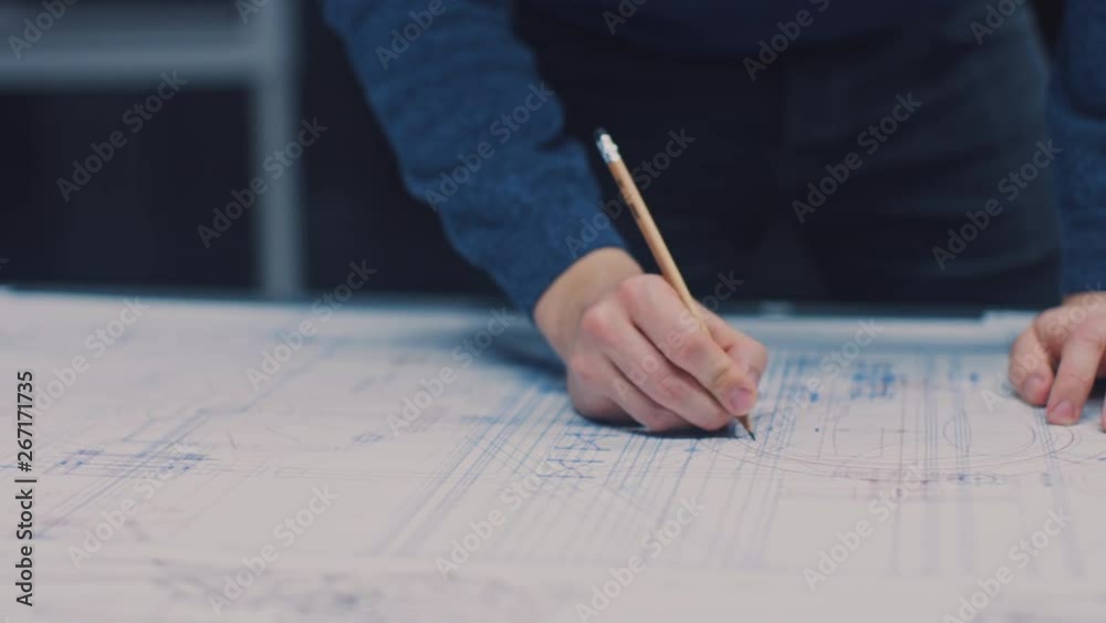 Poster in the dark industrial design engineering facility: male engineer works with blueprints laying on a 