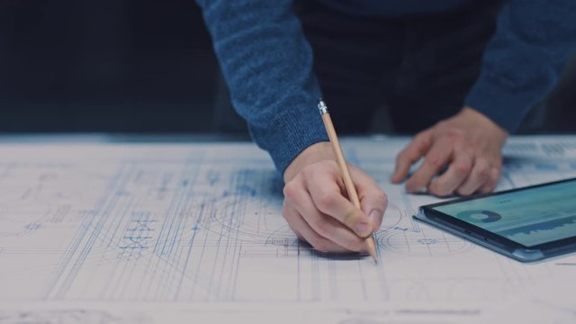 In the Dark Industrial Design Engineering Facility: Male Engineer Works with Blueprints Laying on a Table, Uses Pencil, Ruler and Digital Tablet. On Desktop Multiple Drawings. Focus on Hands