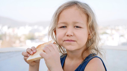 Portrait of little blonde girl is eating toast with butter for breakfast sitting in terrace.