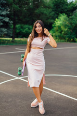 Fototapeta premium Portrait of a smiling charming brunette female holding her skateboard on a basketball court.