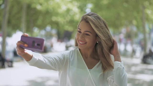 Happy young woman taking selfie outdoor. Beautiful smiling girl taking selfie with smartphone on street at sunny day. Technology concept