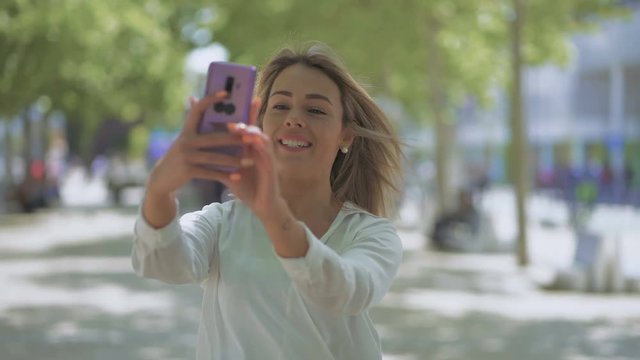 Smiling girl having video chat outdoor. Beautiful happy young woman talking and waving hand during video conversation on street. Communication concept
