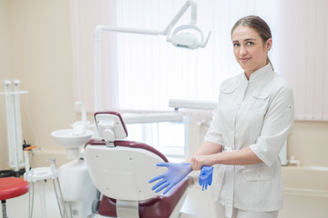 Obraz premium Attractive female doctor in white uniform puts on gloves. Young beautiful female dentist brunette in the office by the chair wearing gloves.