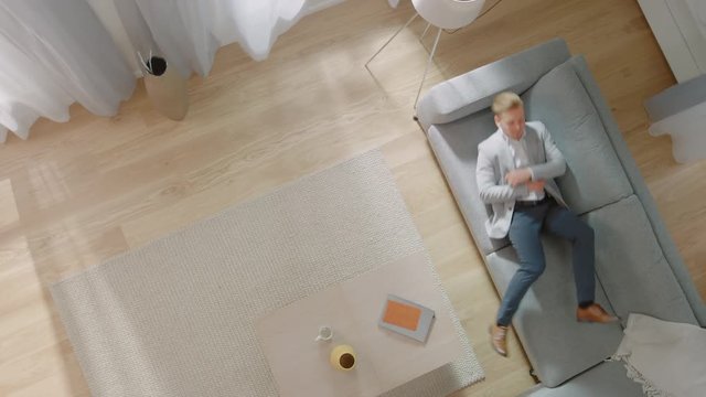 Young Man Comes Home, Dancing and Lying Down on a Sofa. Cozy Living Room with Modern Interior, Grey Sofa and Wooden Flooring. Top View Camera Footage.