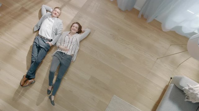 Young Couple are Lying on a Wooden Flooring in an Apartment. They are Happy, Smile and Laugh. Cozy Living Room with Modern Interior, Grey Sofa and Wooden Parquet. Top View Camera Footage.