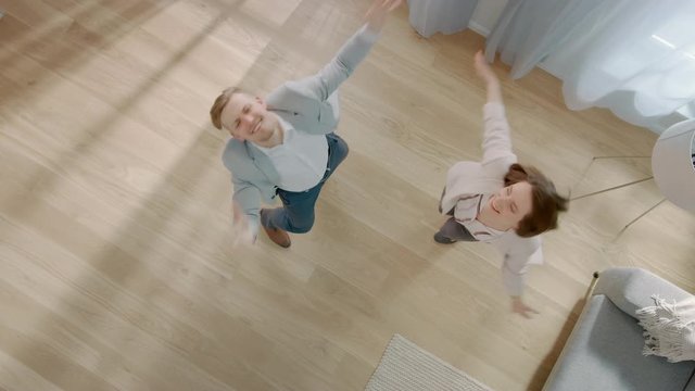 Young Couple is Dancing in an Apartment with Wooden Flooring. They Laugh and Have Fun. Cozy Living Room with Modern Interior, Grey Sofa and Wooden Parquet. Top View Camera Footage.
