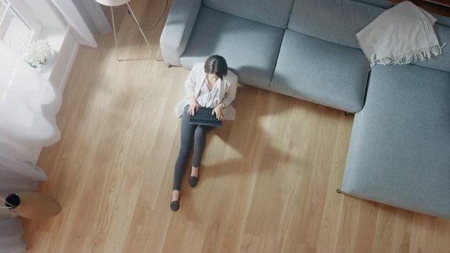 Young Woman is Sitting on a Floor, Working or Studying on a Laptop. Cozy Living Room with Modern Interior, Grey Sofa and Wooden Flooring. Top View Camera Footage.