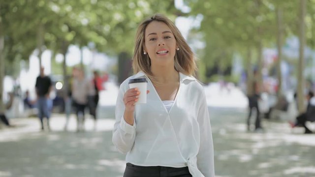 Attractive girl with paper cup walking on street. Beautiful happy young woman holding coffee to go, talking and smiling at camera outdoor. Beverage concept