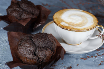 A cup of cappuccino coffee stands on a gray table next to two beautiful chocolate muffins