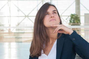 Fototapeta premium Pretty serious Caucasian woman in white blouse and navy blue jacket standing in office corridor, looking aside, pondering, holding tablet in hand. Work concept