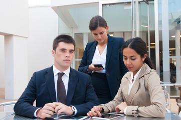 Serious business people using gadgets at desk outdoors. Business man and women, browsing on tablet computers and smartphone, standing and sitting at cafe table. Communication concept. Front view.