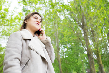 Smiling pretty young woman calling on smartphone in park. Lady wearing jacket, talking on mobile phone and standing with green trees in background. Communication and nature concept.