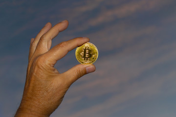 A bitcoin coin in gold is held by one hand, in the background a blue sky with clouds and beautiful...