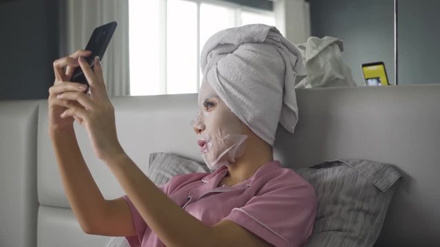 Happy young woman with facial mask taking selfie photo using a mobile phone in the bedroom at home. Shot in 4k resolution